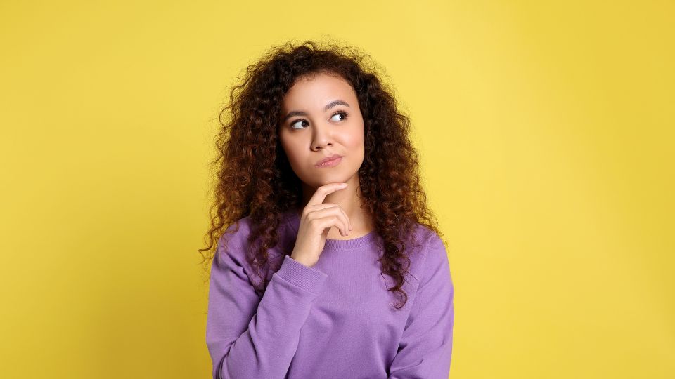 Une femme avec des cheveux bouclés portant un pull violet, réfléchissant avec une main sur le menton sur un fond jaune.