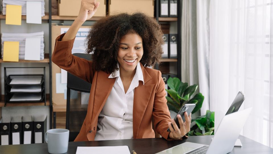 Une femme souriante, assise à son bureau, levant le poing en signe de réussite tout en regardant son téléphone portable.