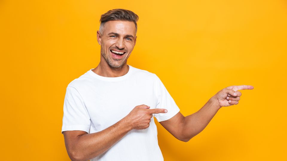 Un homme souriant portant un t-shirt blanc, pointant vers la droite avec ses deux mains sur un fond jaune.