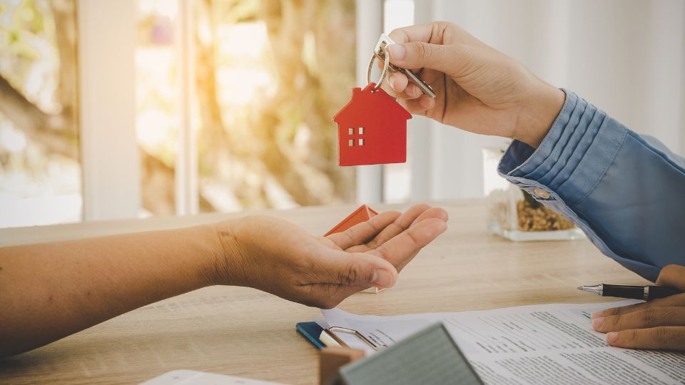 Une personne remet des clés avec un porte-clés en forme de maison à une autre personne au-dessus d'un bureau avec des documents.