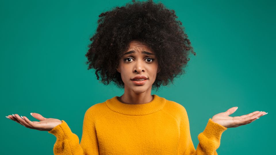 Une femme avec des cheveux bouclés et un pull jaune, levant les bras avec une expression incertaine sur un fond vert.