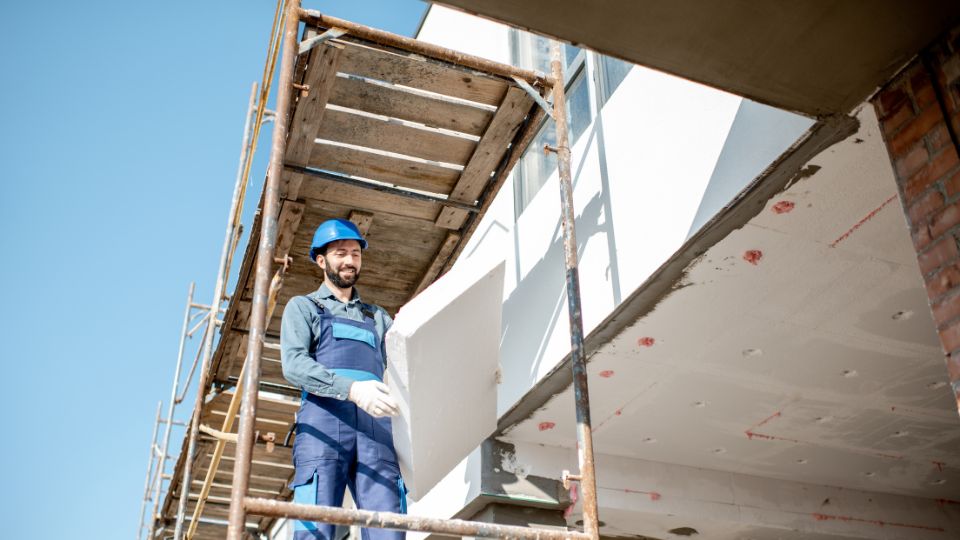 Un ouvrier portant un casque bleu et une combinaison de travail, tenant un panneau d'isolation thermique sur un échafaudage.