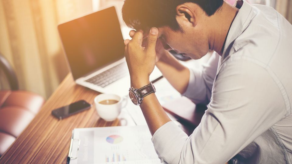 Un homme assis à son bureau, tenant sa tête entre ses mains avec des graphiques financiers et un ordinateur portable à côté de lui.