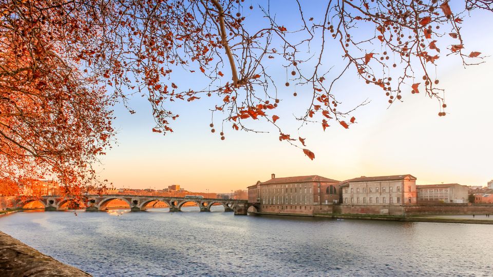 Vue du Pont Neuf à Toulouse au coucher du soleil, encadrée par des branches d'arbre aux feuilles rouges.