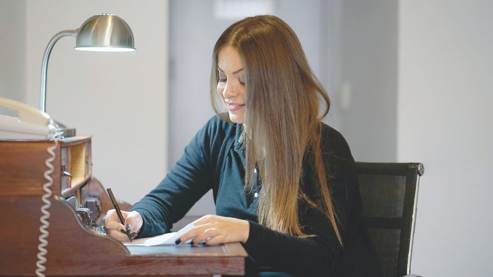 Une femme souriante assise à un bureau en bois, rédigeant une lettre sous une lampe de bureau.