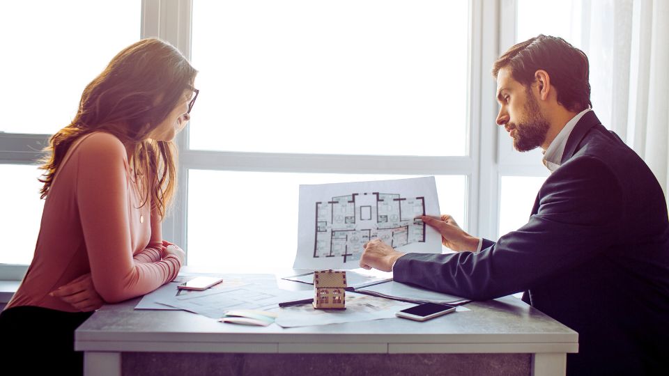 Un agent immobilier montre des plans d'étage à une cliente dans un bureau bien éclairé.