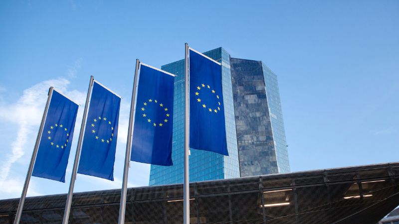 Quatre drapeaux de l'Union Européenne flottant devant un bâtiment moderne en verre sous un ciel bleu clair.