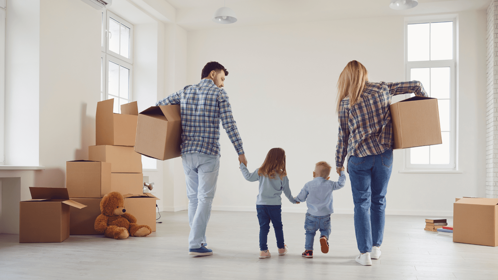 Une famille de quatre personnes portant des cartons et se tenant par la main en entrant dans une nouvelle maison.