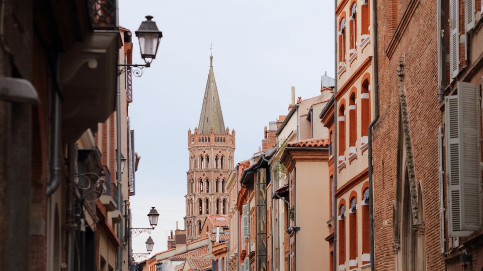 Rue étroite typique de Toulouse avec des bâtiments en briques rouges et une vue sur un clocher au loin.